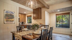 Un coin repas avec une table en bois dressée pour quatre personnes, entourée de chaises en bois foncé. Un grand luminaire suspendu se trouve au-dessus de la table, et une affiche au mur représente un paysage pittoresque.