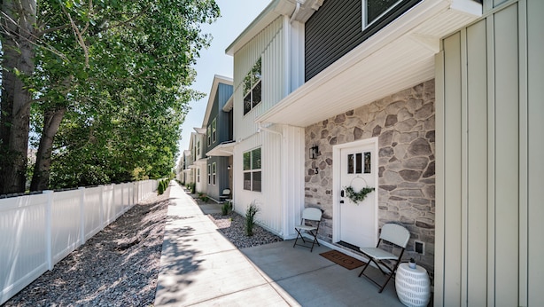 A residential property with a stone facade, a white picket fence, and a small porch area with two chairs and a potted plant. - Brand New Townhome in proximity to the Hospital, Mall, Restaurants, and Parks. (Idaho Falls)