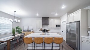 A modern kitchen with a central island, bar stools, white cabinetry, stainless steel appliances, and a dining area with a bench and chairs. - Brand New Townhome in proximity to the Hospital, Mall, Restaurants, and Parks. (Idaho Falls)