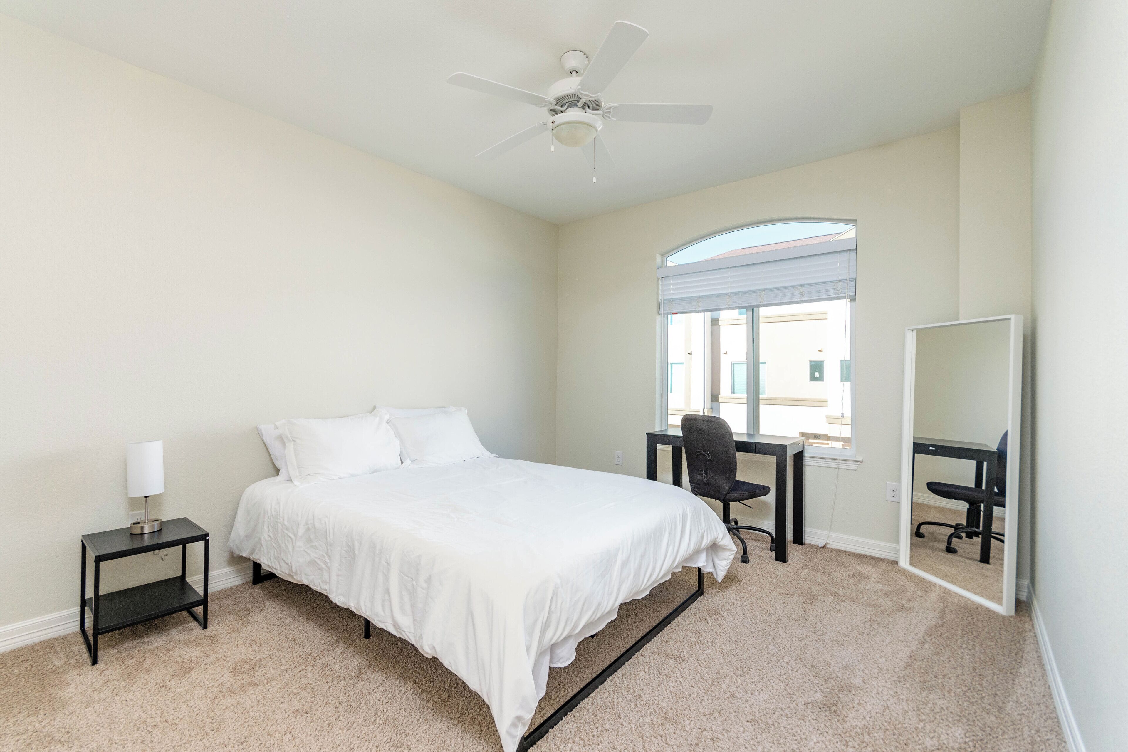 A bedroom with a bed, a desk, a chair, a lamp, a ceiling fan, and a window.