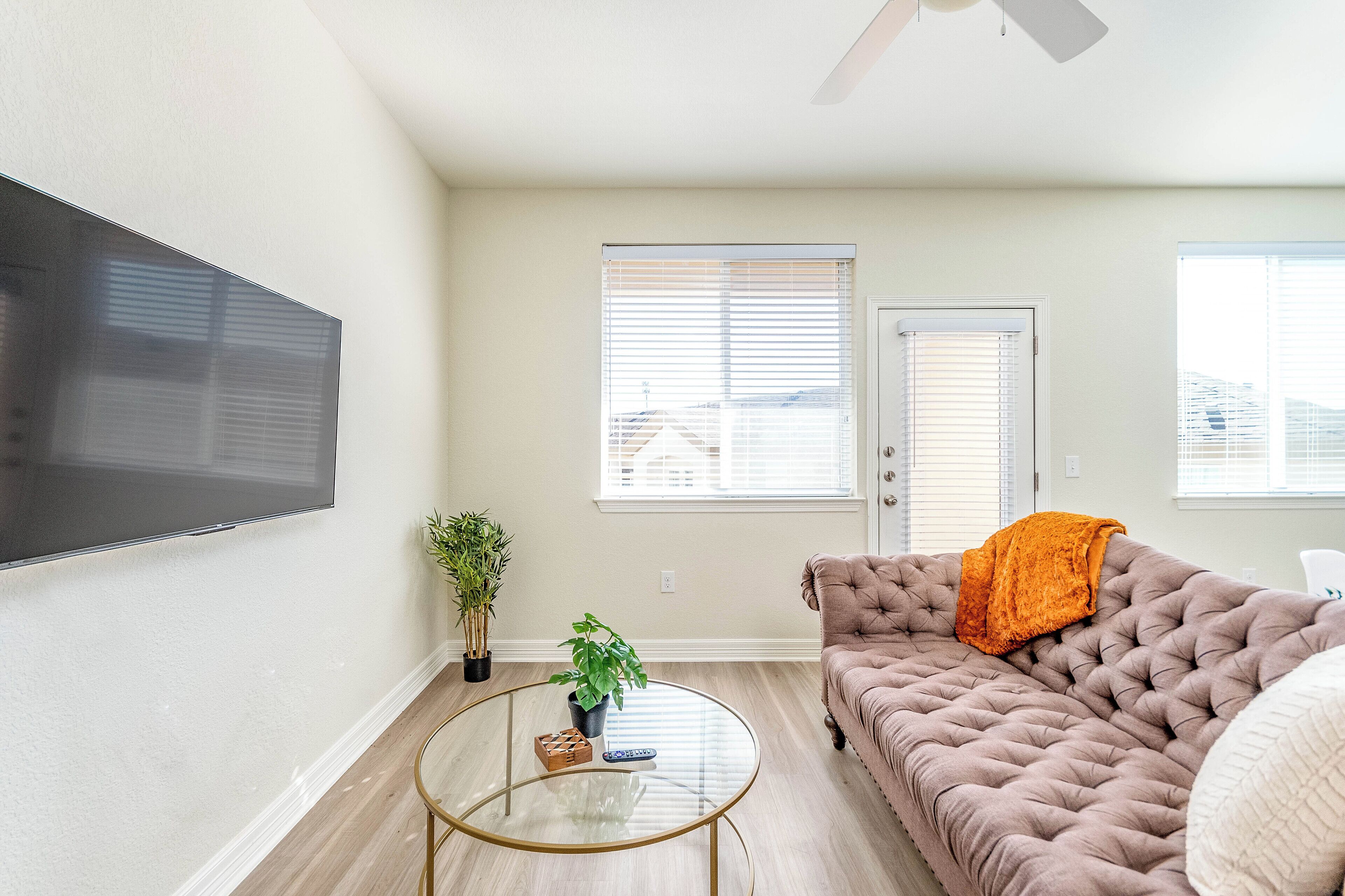 Un salon avec un téléviseur à écran plat fixé au mur, un canapé capitonné agrémenté d’un plaid orange, une table basse en verre et une plante en pot.