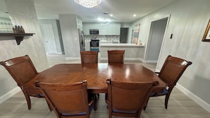 Un comedor con una mesa y sillas de madera, una araña y una cocina al fondo.