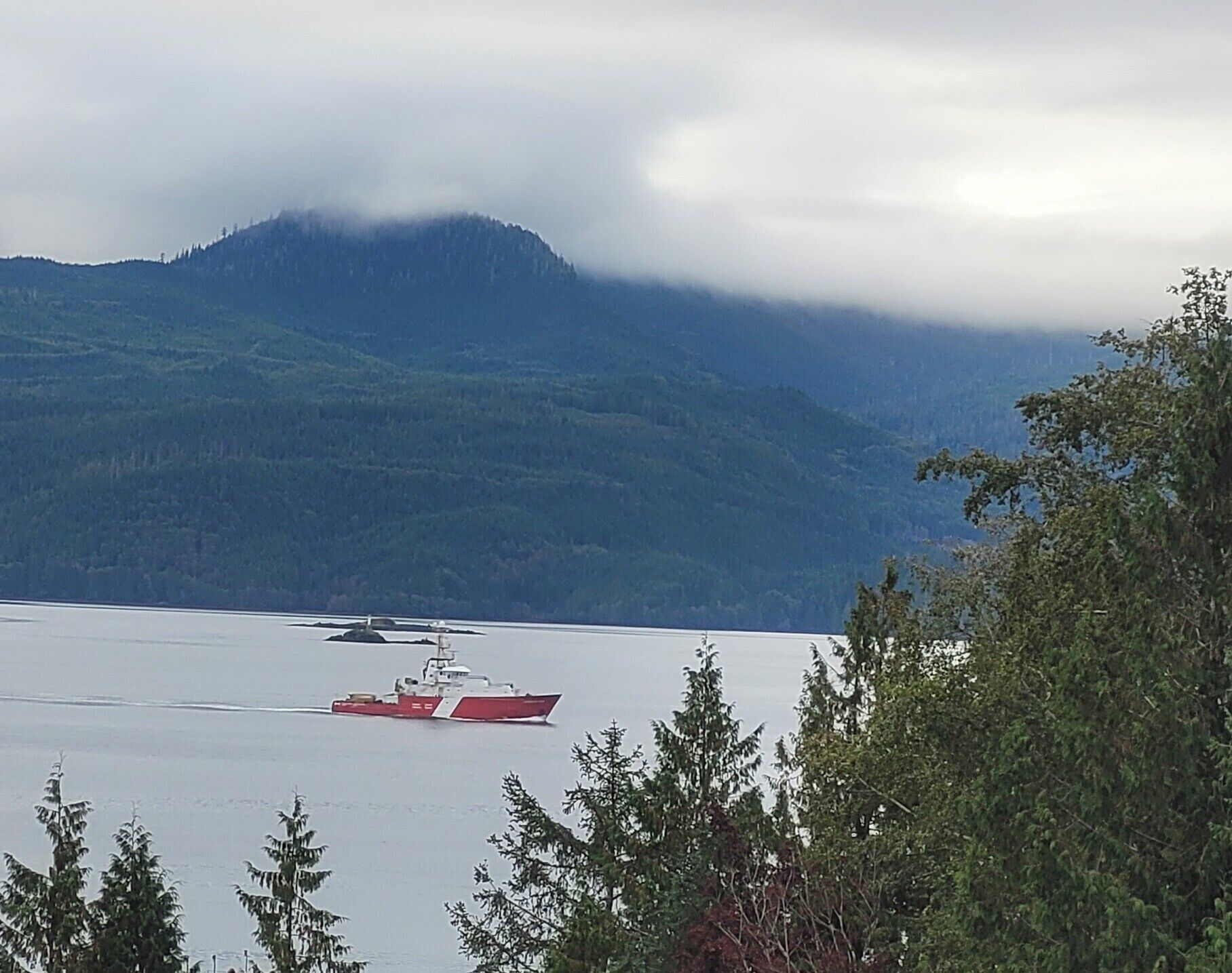 Une vaste étendue d’eau, avec un navire rouge visible au loin et une chaîne de montagnes partiellement masquée par les nuages.
