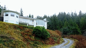 Ett hus med ett sluttande tak och en balkong, omgivet av ett skogsområde och en slingrande väg.