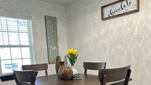 Een eetruimte met een houten tafel en stoelen, een vaas met gele bloemen en een bordje aan de muur.