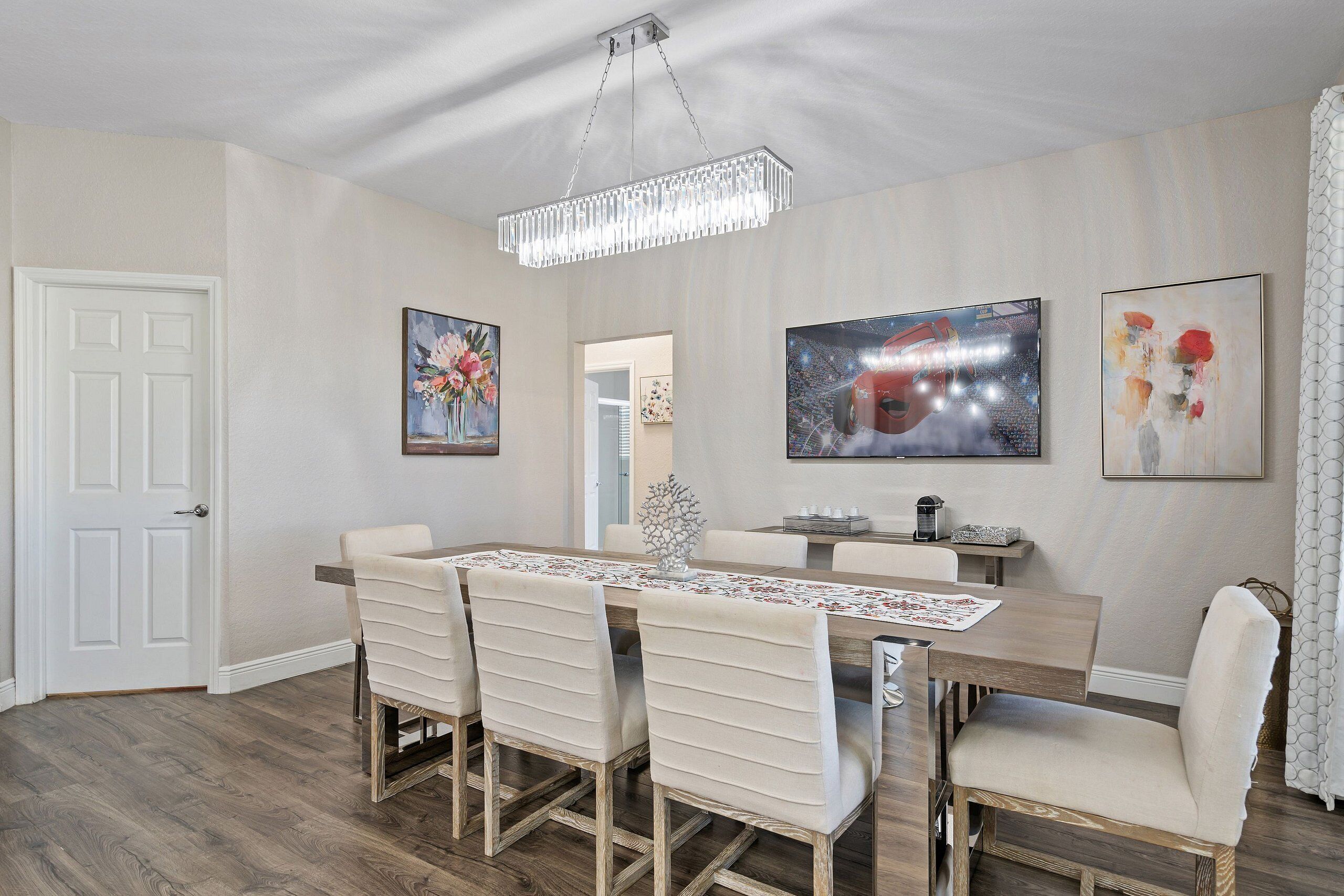 A dining area with a wooden table, white upholstered chairs, a chandelier, and artwork on the walls.