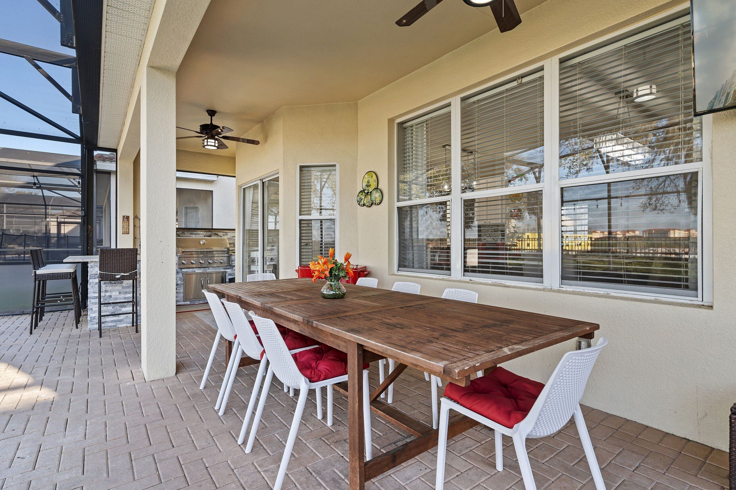 An outdoor patio with a wooden table and white chairs, large windows with blinds, and a ceiling fan.