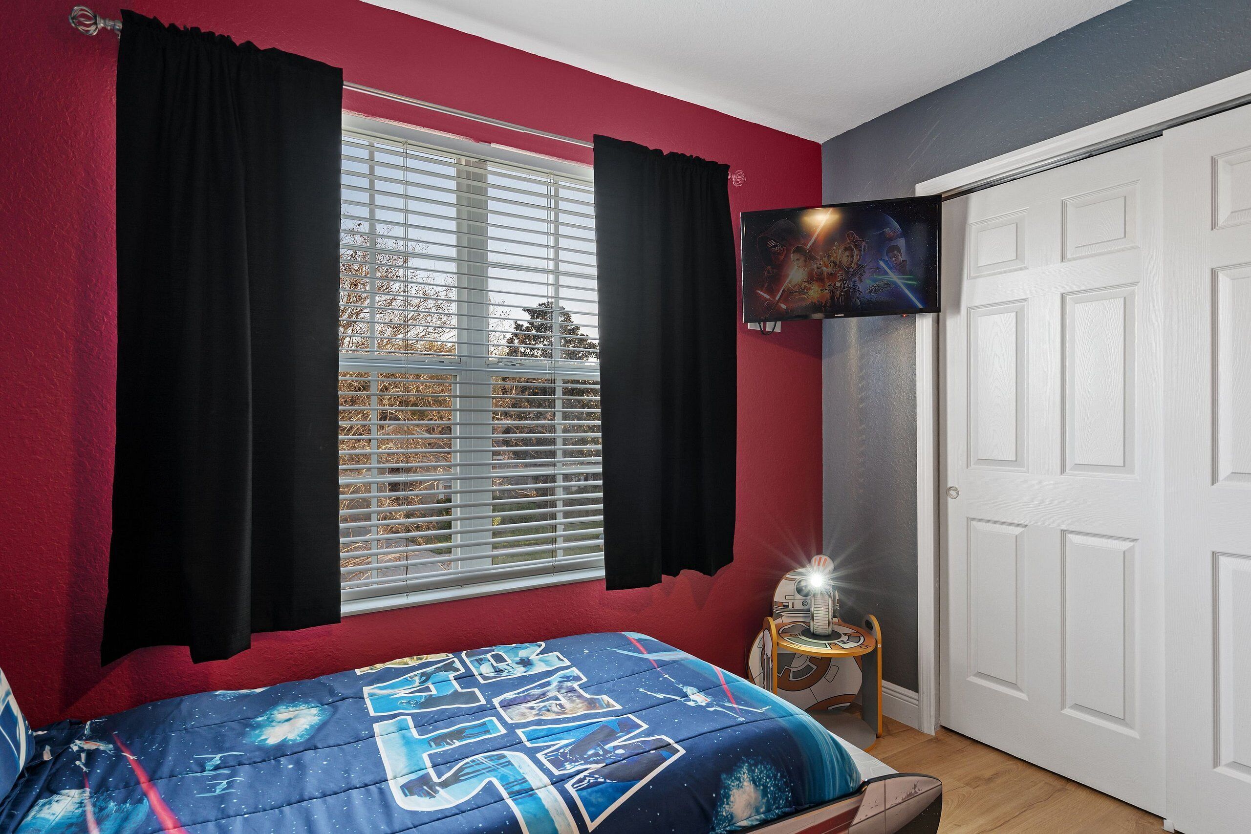 Une chambre à coucher avec un lit, une fenêtre avec des rideaux, un téléviseur fixé au mur et une porte.