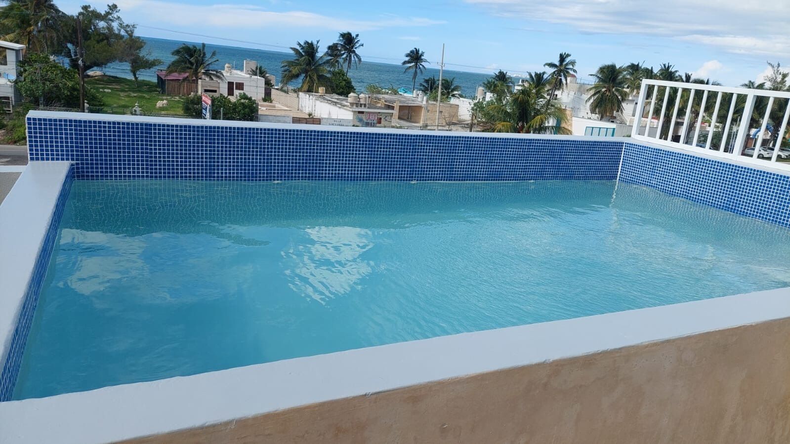Une piscine aux carreaux bleus, entourée d’une terrasse blanche et de palmiers.
