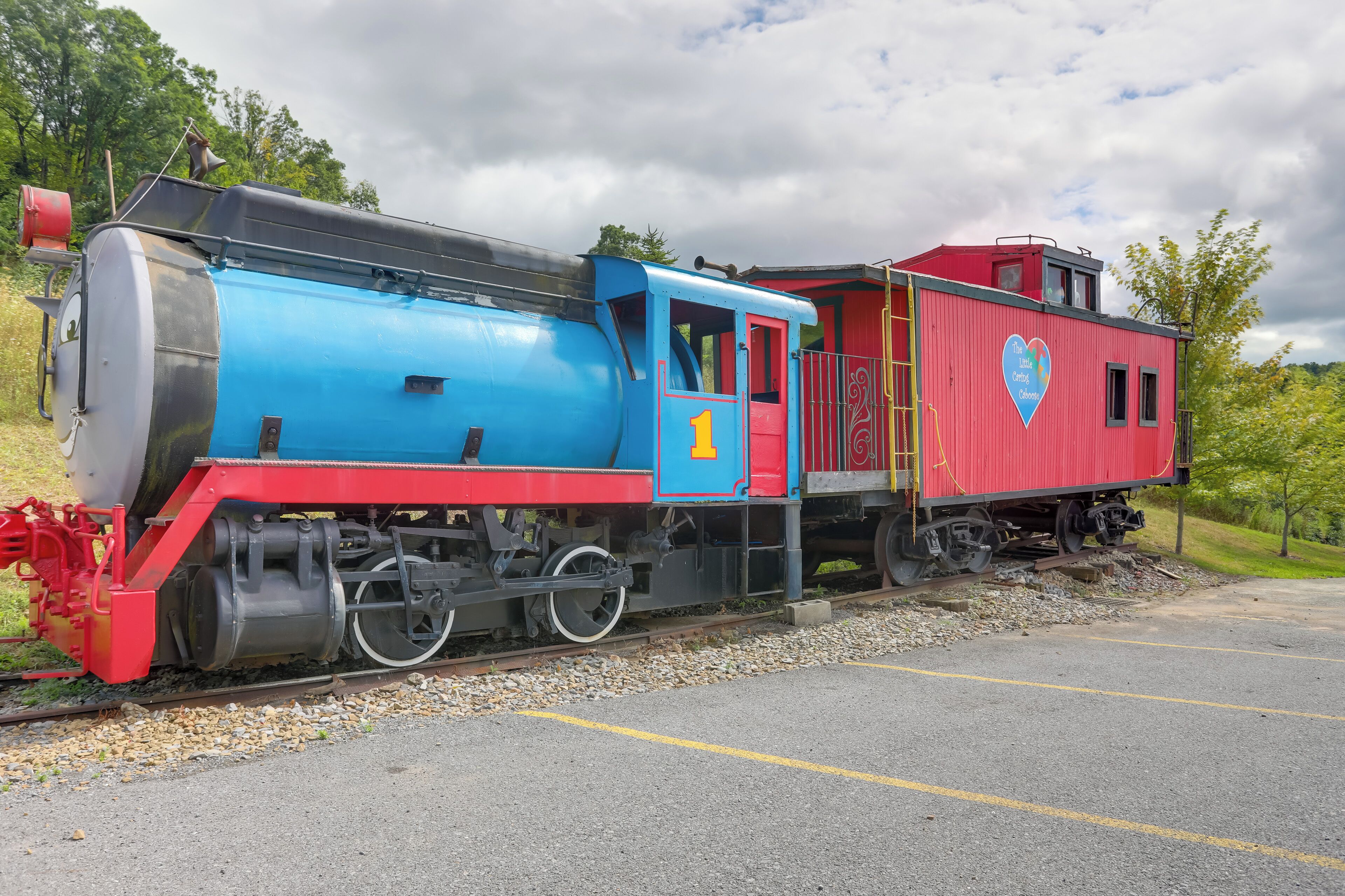 Une locomotive à vapeur d’époque, avec un wagon de queue rouge et bleu, est garée sur une voie ferrée dont le ballast est constitué de gravier, et une zone pavée se trouve à côté.