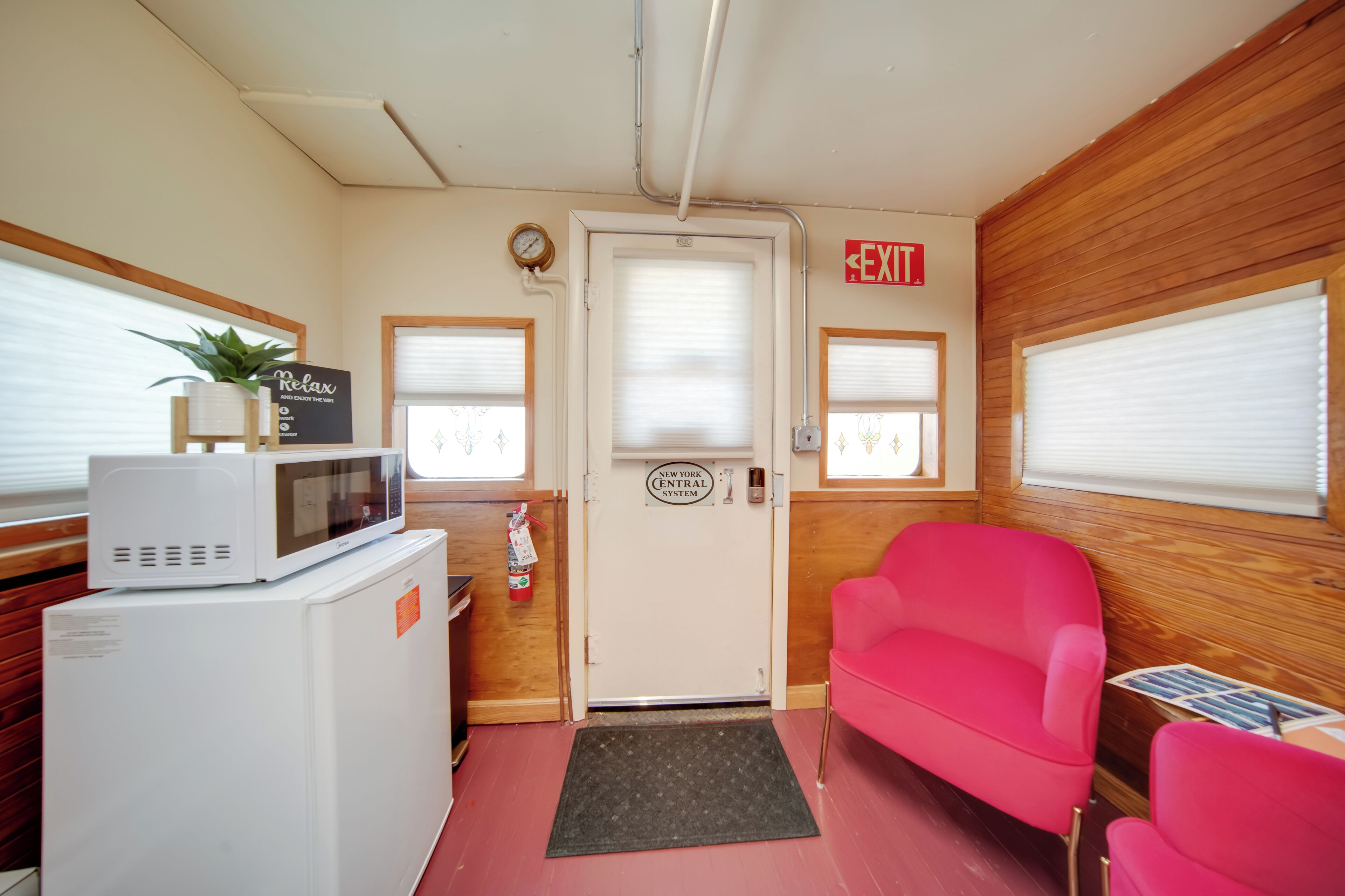A room with a microwave, a small refrigerator, a red chair, a wooden wall, and a door with an exit sign.
