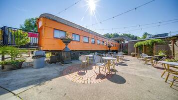 Un espacio de patio al aire libre con mesas y sillas, un hogar de fuego de ladrillo y un vagón de tren naranja antiguo.