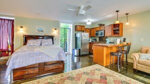 A bedroom with a large bed, a wooden dresser, and a nightstand. There is a kitchen area with wooden cabinets, a refrigerator, and a microwave.