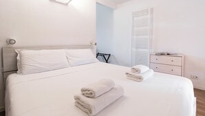 Una cama bien hecha con sábanas blancas y dos toallas dobladas. Hay una mesita de noche con una lámpara y un cajón pequeño. La habitación tiene una pared de acento azul claro y un techo blanco.
