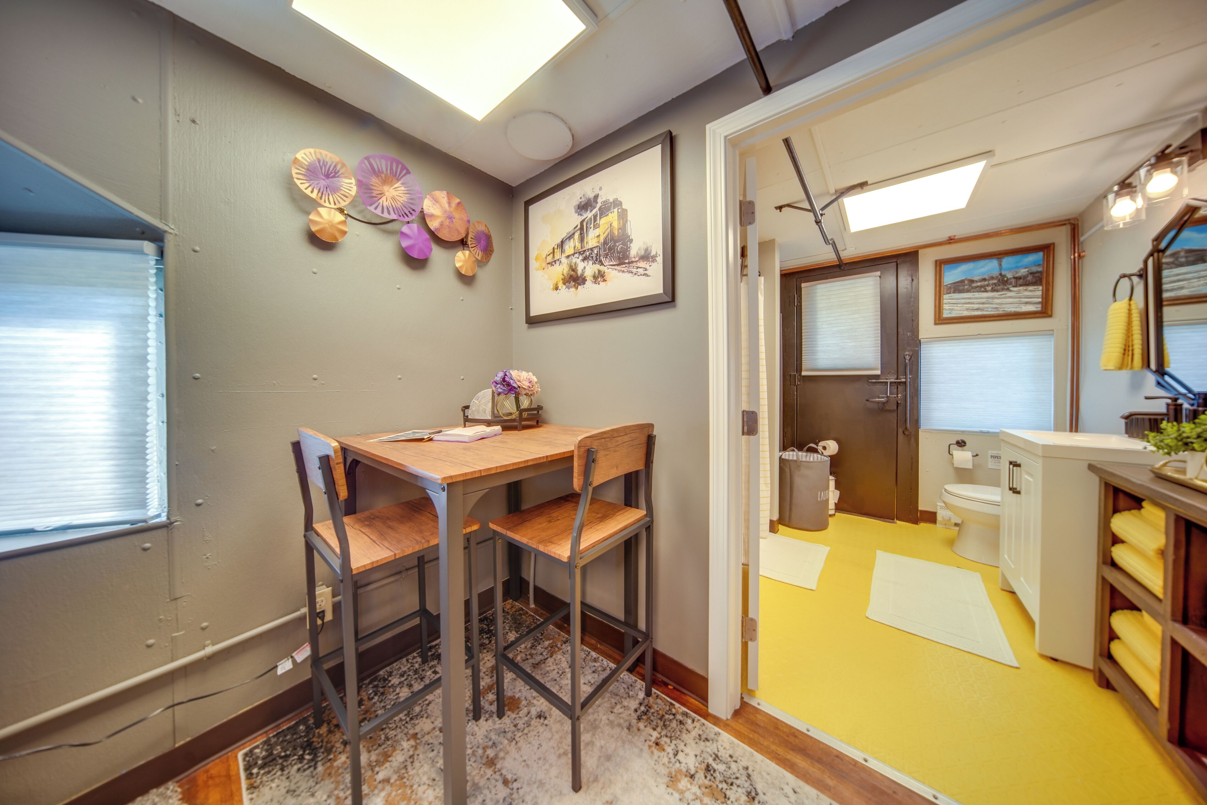 A room with a wooden desk, two chairs, a window with blinds, a framed picture on the wall, and a bathroom with a yellow floor.