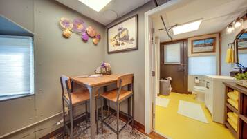 A room with a wooden desk, two chairs, a window with blinds, a framed picture on the wall, and a bathroom with a yellow floor.