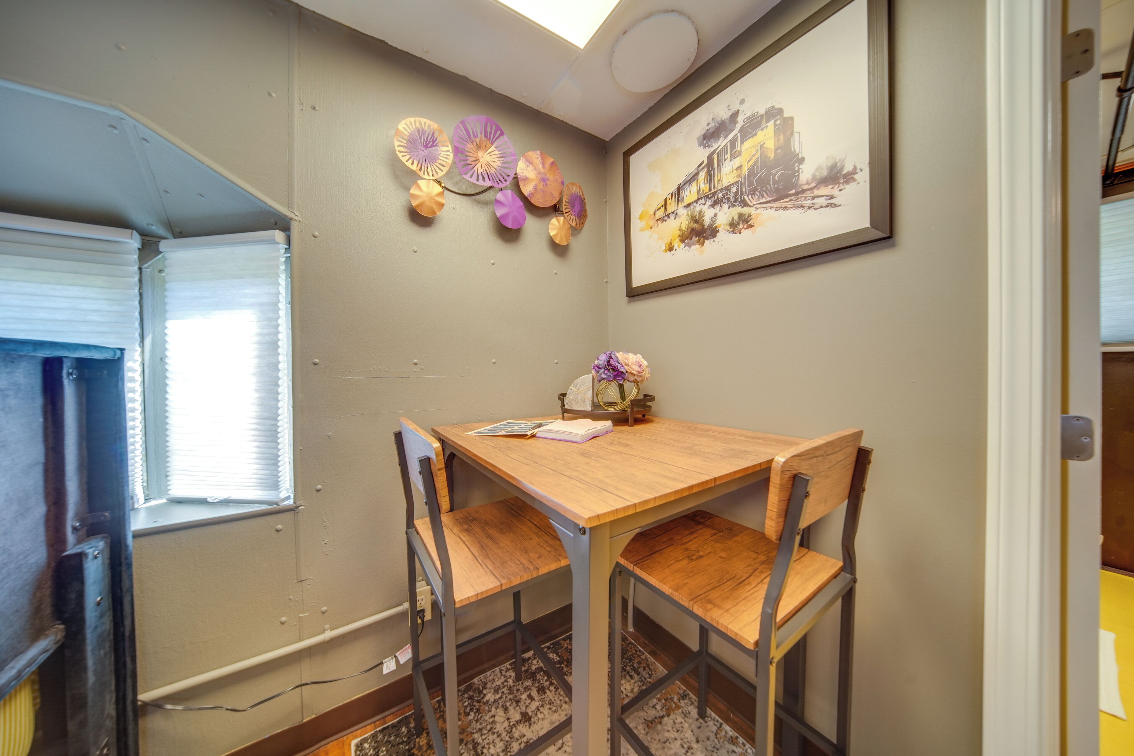 Un comedor con una mesa de madera y sillas metálicas, una ventana con persianas, y decoración en las paredes, incluyendo abanicos de papel y un cuadro enmarcado.