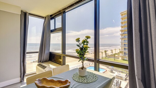 A room with a view of the beach, a table with a bowl and a potted plant, and a large window with curtains. - Beachfront Studio: Walk to Daytona Fun! (Daytona Beach Shores)