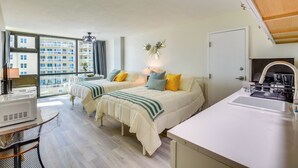 A hotel room with two beds, a kitchenette, a large window with a view of buildings, and a ceiling fan. - Beachfront Studio: Walk to Daytona Fun! (Daytona Beach Shores)