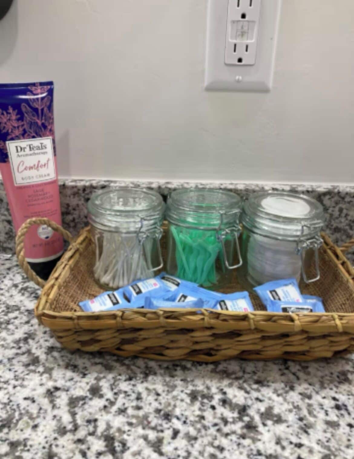 A wicker basket on a granite countertop containing green sachets, a pink bottle, and blue packets.