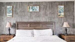 Un dormitorio con cabecera de madera, ropa de cama blanca y dos mesitas de noche con lámparas. Hay tres cuadros enmarcados en la pared.