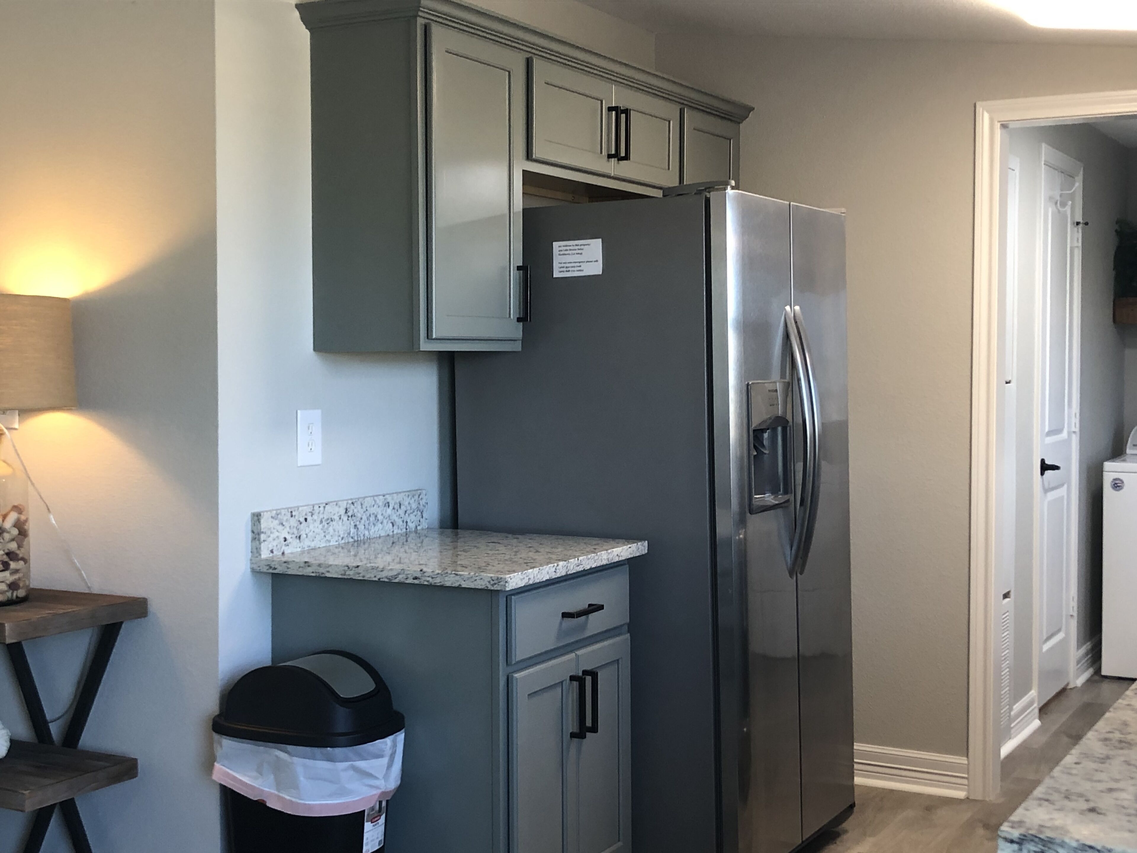 A kitchen with a stainless steel refrigerator, grey cabinets, a granite countertop, and a trash can.