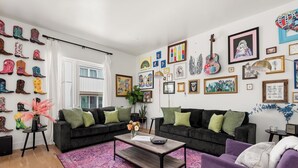 A living room with two dark sofas, a coffee table, a purple armchair, a pink rug, and a wall adorned with various framed artworks and a collection of colorful boots.