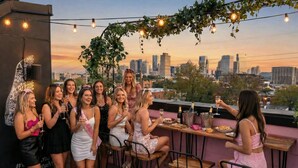 A group of people on a rooftop terrace with a city skyline view, string lights, and a bar area with drinks.