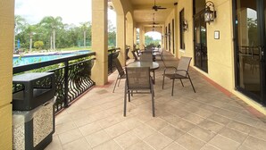Un patio al aire libre con mesas y sillas, una piscina al fondo y vistas a palmeras.
