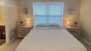 Un dormitorio con una cama grande, dos mesitas de noche con lámparas, una ventana con persianas y una puerta que da a un baño.
