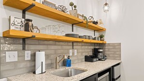 A kitchen with a granite countertop, stainless steel sink, and black appliances. There are wooden shelves above the countertop holding various kitchen items. - Birdie Suite by The Inn & Spa at Cedar Falls (Logan)