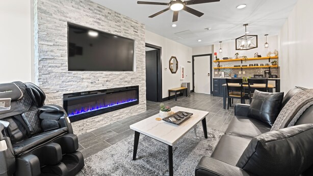 A modern living room with a stone wall, a flat-screen TV, a fireplace, a massage chair, a coffee table, and a dining area with a bar. - Birdie Suite by The Inn & Spa at Cedar Falls (Logan)