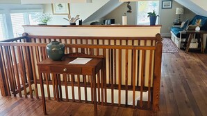 Una habitación con piso de madera, una mesa de madera con un florero, un banco y vista a un balcón con barandilla.