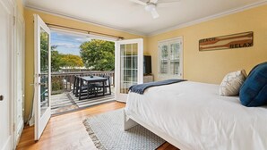 A bedroom with a bed, a ceiling fan, a view through an open door to a balcony with a table and chairs, and a 'BEACH RELAX' sign on the wall. - Historic Seaside Beach Home | Deck, Hot Tub, Games (Seaside)