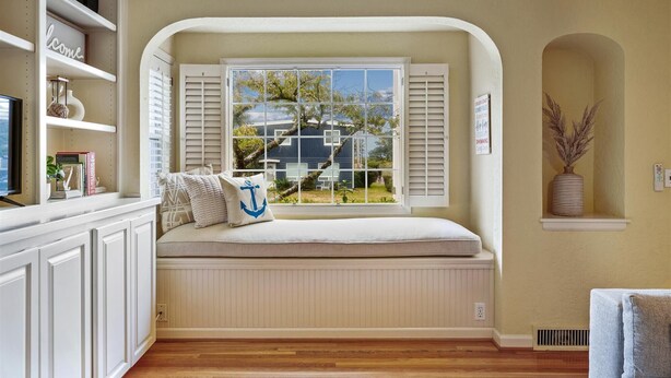 A cozy room with a built-in bench under a large window, a television on the left, and a small table with decorative items. - Historic Seaside Beach Home | Deck, Hot Tub, Games (Seaside)
