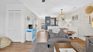 Una sala moderna con un sofá, una mesa de centro y un ventilador de techo. Se observa un área de cocina con una encimera y armarios al fondo.