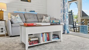 Una sala de estar con un sofá gris, una mesa de centro blanca con libros, una lámpara y vistas al exterior a través de una ventana con cortinas azules.