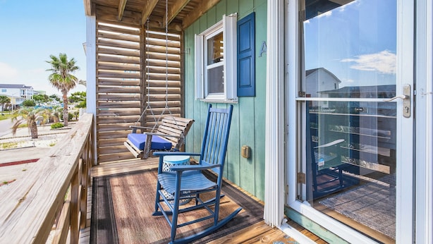 Una terraza de madera con una mecedora y una mesa, una puerta corrediza de vidrio y vista a la playa.