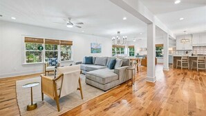 A spacious living area with a grey sectional sofa, a small round table, and a chair. There is a kitchen area with bar stools and a chandelier. - Now Offering up to 25% OFF Fall 2026, New in 2026 , Steps to Beach, Pool, Free Golf, Dogs OK (Hilton Head Island)