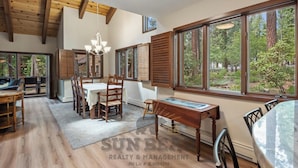 Uma sala de jantar ampla, com mesa e cadeiras de madeira, grandes janelas com vista para a floresta e um lustre pendurado no teto.