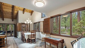 Une salle à manger spacieuse avec une table et des chaises en bois, de grandes fenêtres donnant sur la forêt et un lustre suspendu au plafond.