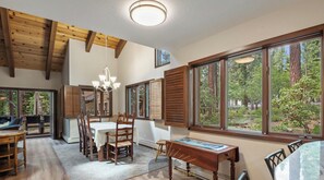 Une salle à manger spacieuse avec une table et des chaises en bois, de grandes fenêtres donnant sur la forêt et un lustre suspendu au plafond.