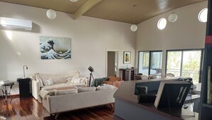 A modern living room with a sofa, a coffee table, and a dining area with a table and chairs. There is a bookshelf with books and a painting on the wall. - Culburra Beach Lake House (Culburra Beach)
