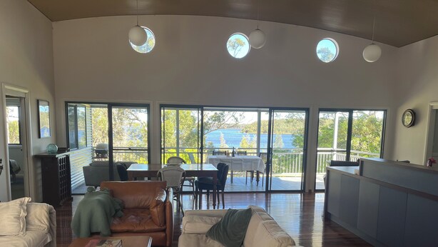 A spacious living area with a large sliding glass door leading to a balcony with a view of the sea. There is a dining area with a table and chairs, and a kitchen area with a counter and cabinets. - Culburra Beach Lake House (Culburra Beach)