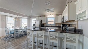 A modern kitchen with white cabinetry, a central island with bar stools, a ceiling fan, and a dining area with a glass table and chairs. - Sea Time - Corolla, Ocean Sands, Oceanside Retreat, 280 Yds to Beach, Close to Golf and Shopping (Corolla)