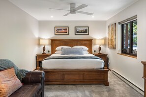 A bedroom with a wooden bed, two bedside tables with lamps, a ceiling fan, framed pictures on the wall, and a window with a view.