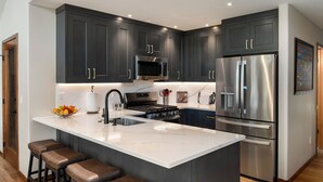 Una cocina moderna con armarios oscuros, electrodomésticos de acero inoxidable, encimera de mármol y taburetes de bar.