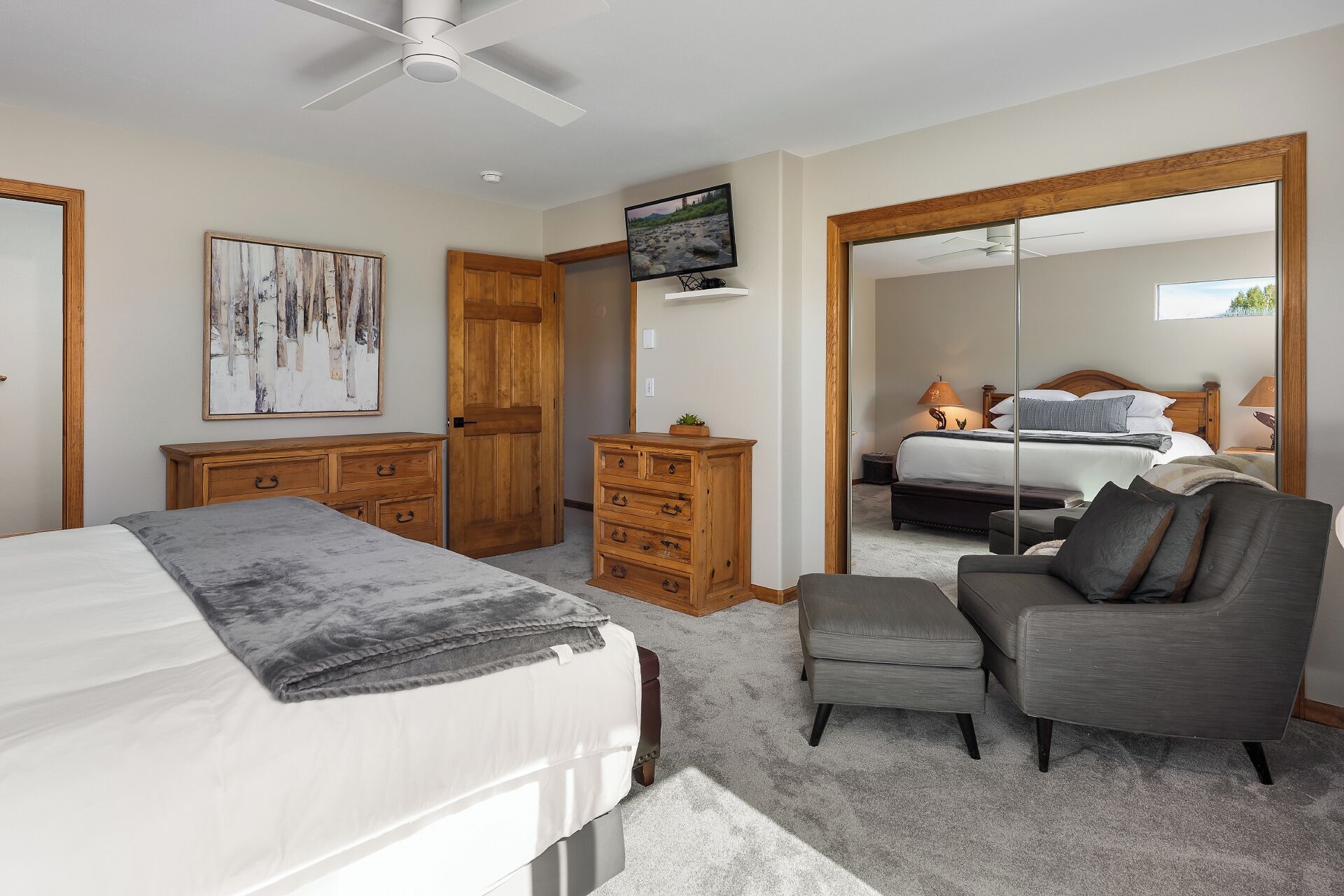 A bedroom with a bed, a grey armchair, a wooden dresser, a television, and a large mirror.