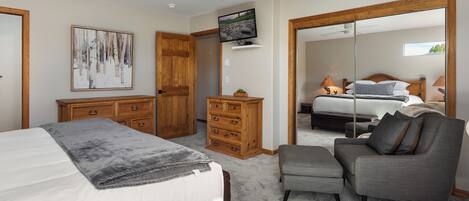 A bedroom with a bed, a grey armchair, a wooden dresser, a television, and a large mirror.