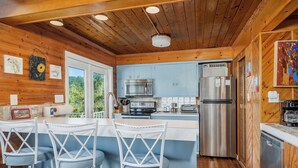 A kitchen with wooden walls, a bar area with white chairs, stainless steel appliances, and a view of greenery outside. - The Lazy Lake House on Center Hill (Sparta)